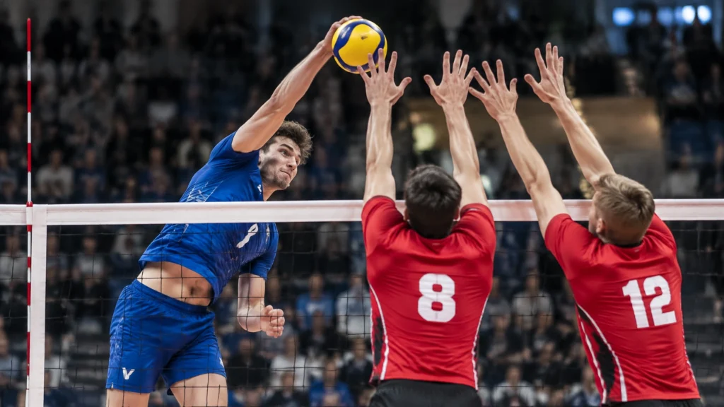 Ein Spieler in blauem Trikot schlägt den Ball über das Netz, zwei Spieler in roten Trikots springen zum Block.